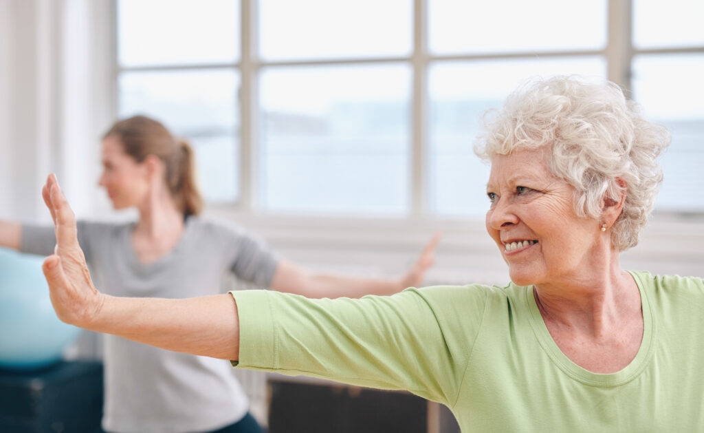 An elderly person is exercising in a bright room, smiling as they stretch their arms out. Another person is visible in the background.