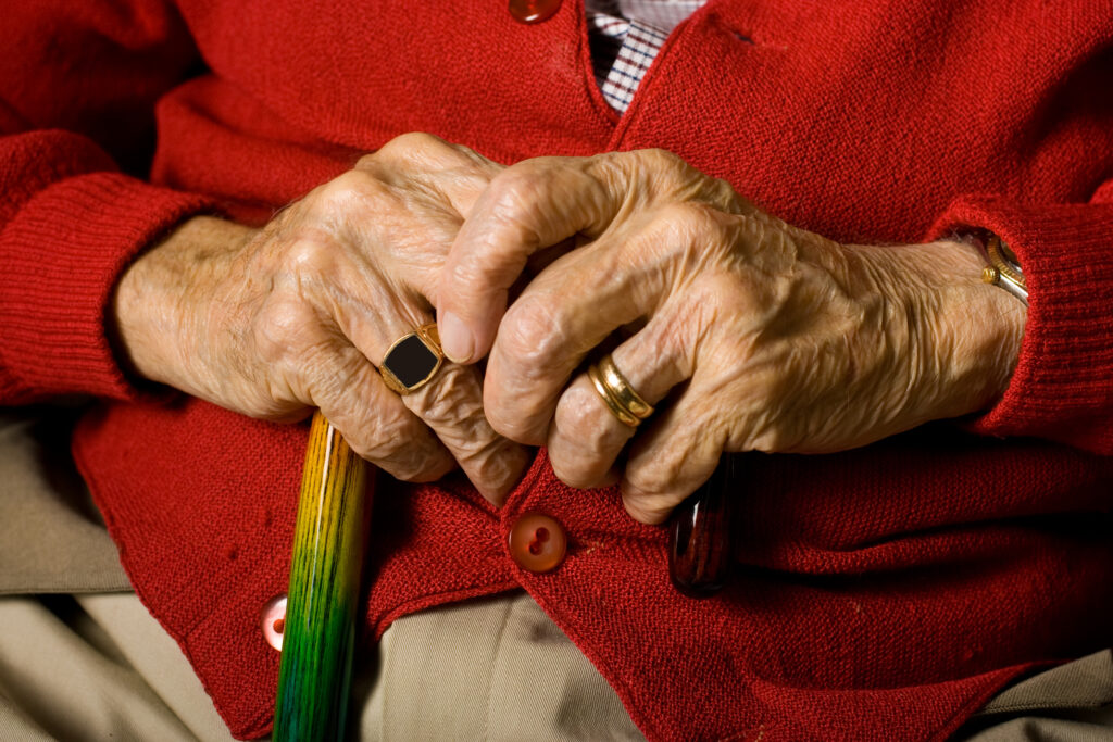 An elderly person's hands rest on their lap, one holding a cane. They wear a red cardigan, a gold ring, and have signs of aged skin.