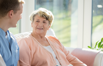 A person and an elderly person are talking and smiling indoors. Sunlight floods the room, suggesting a comforting and warm atmosphere. They seem to be enjoying a pleasant interaction.