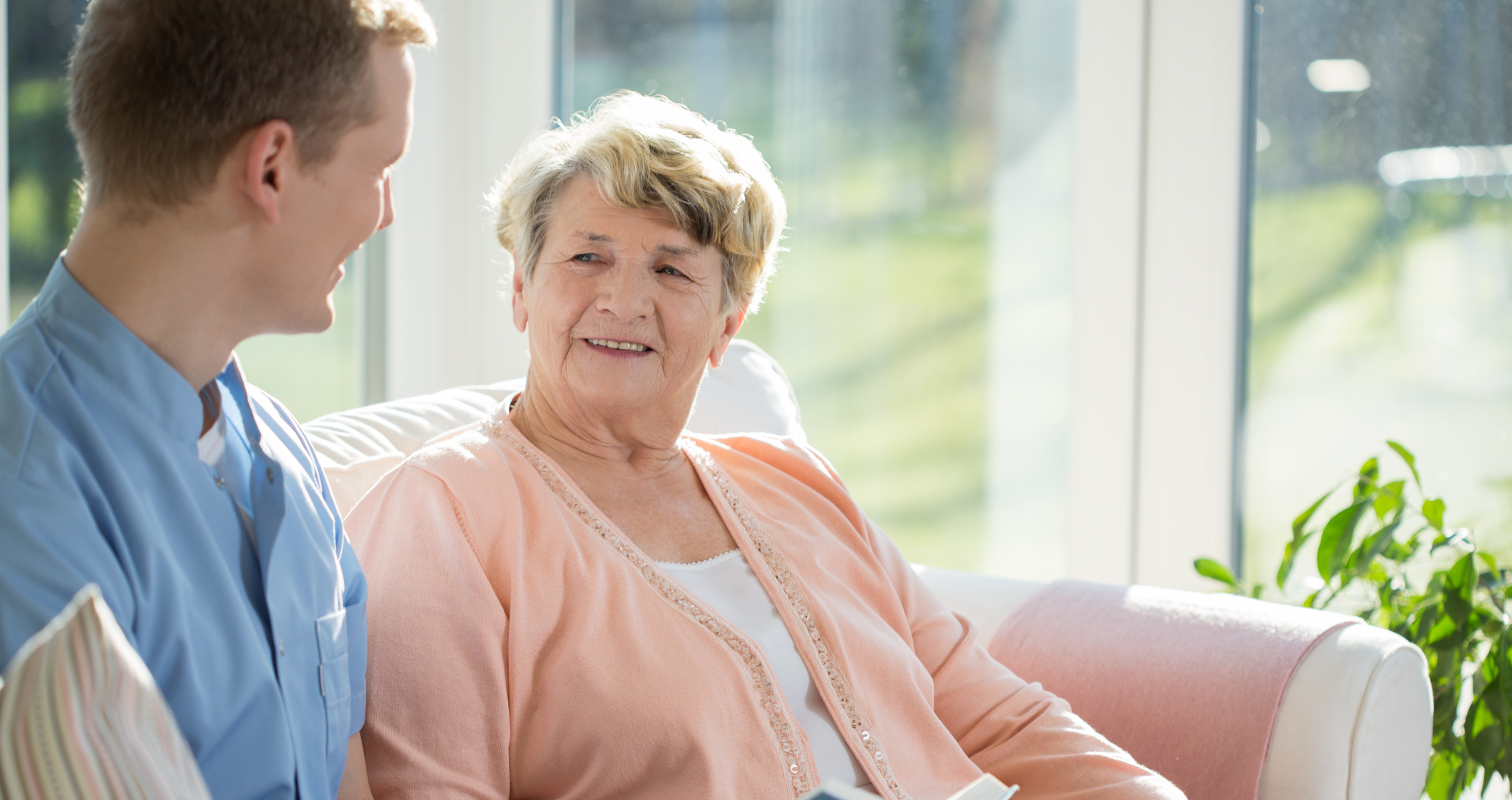 A person and an elderly person are talking and smiling indoors. Sunlight floods the room, suggesting a comforting and warm atmosphere. They seem to be enjoying a pleasant interaction.