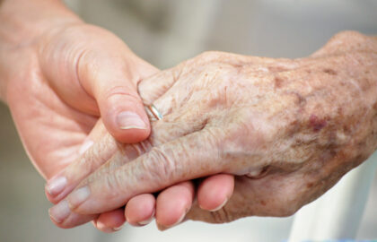 Two people holding hands gently, one with younger skin and the other with older, aged skin featuring prominent veins and age spots, symbolizing care or support.