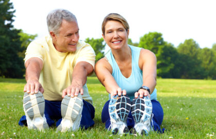 Two smiling adults are stretching in a sunny park, touching their toes while sitting on the grass. They're wearing casual athletic clothing and sneakers.