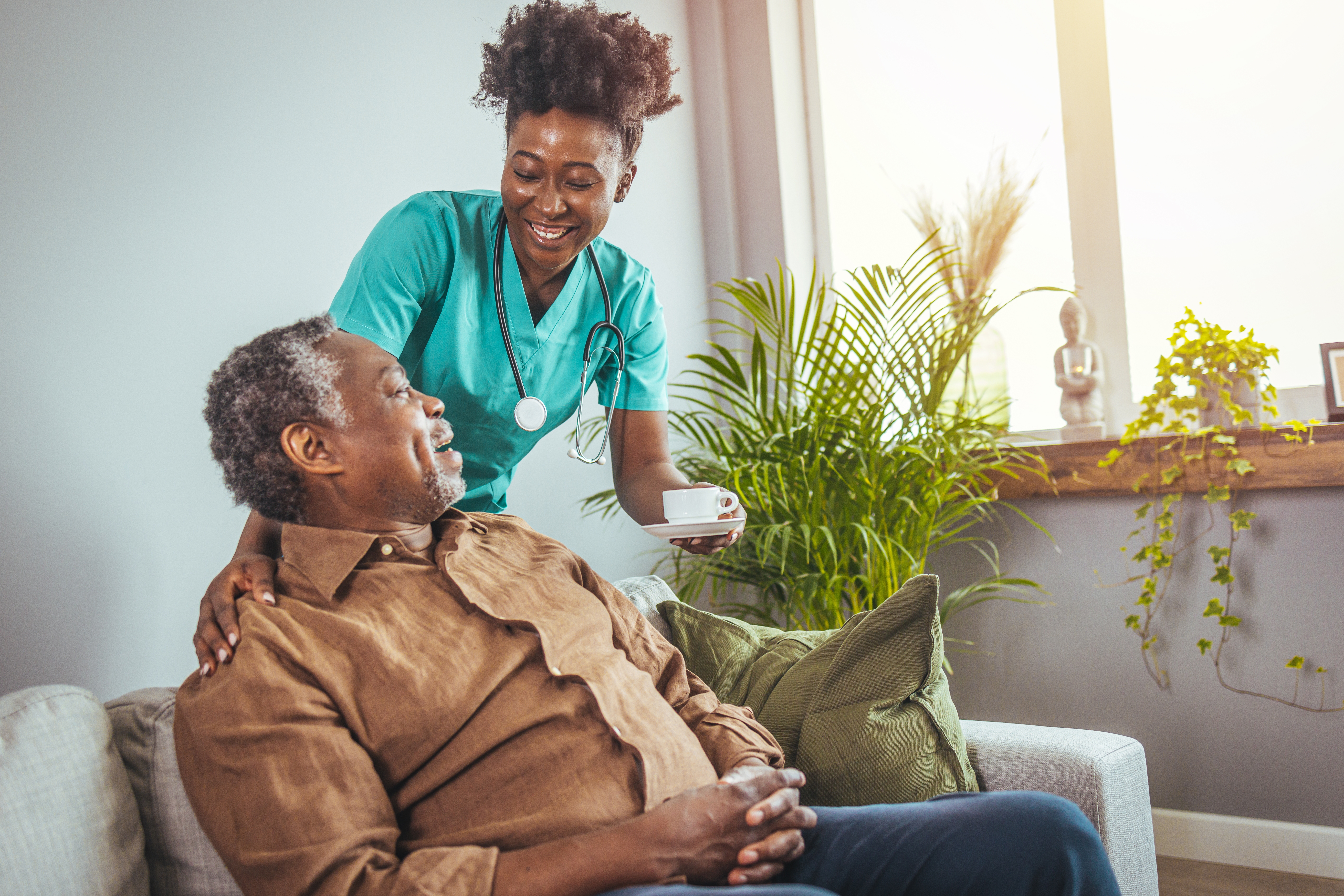 Older man sitting on his couch with a supportive live-in care attendant handing him a cup of tea