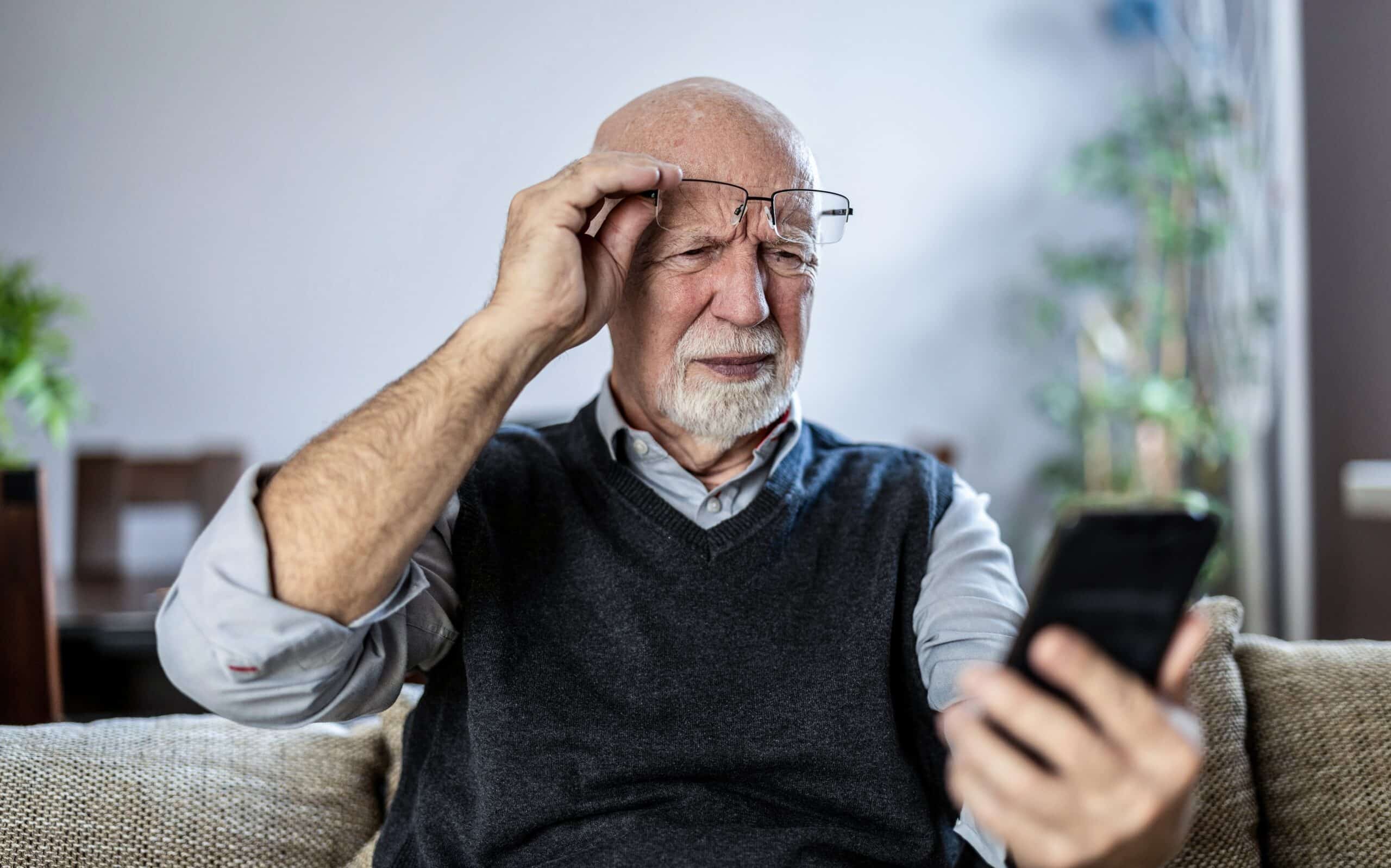 Senior man looking at cell phone while lifting glasses with confused look