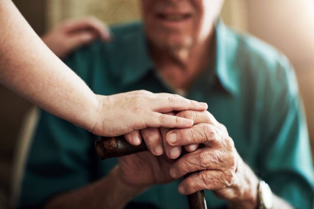 Caregiver offering comfort by placing their hand on the hands of a senior man who is holding a cane