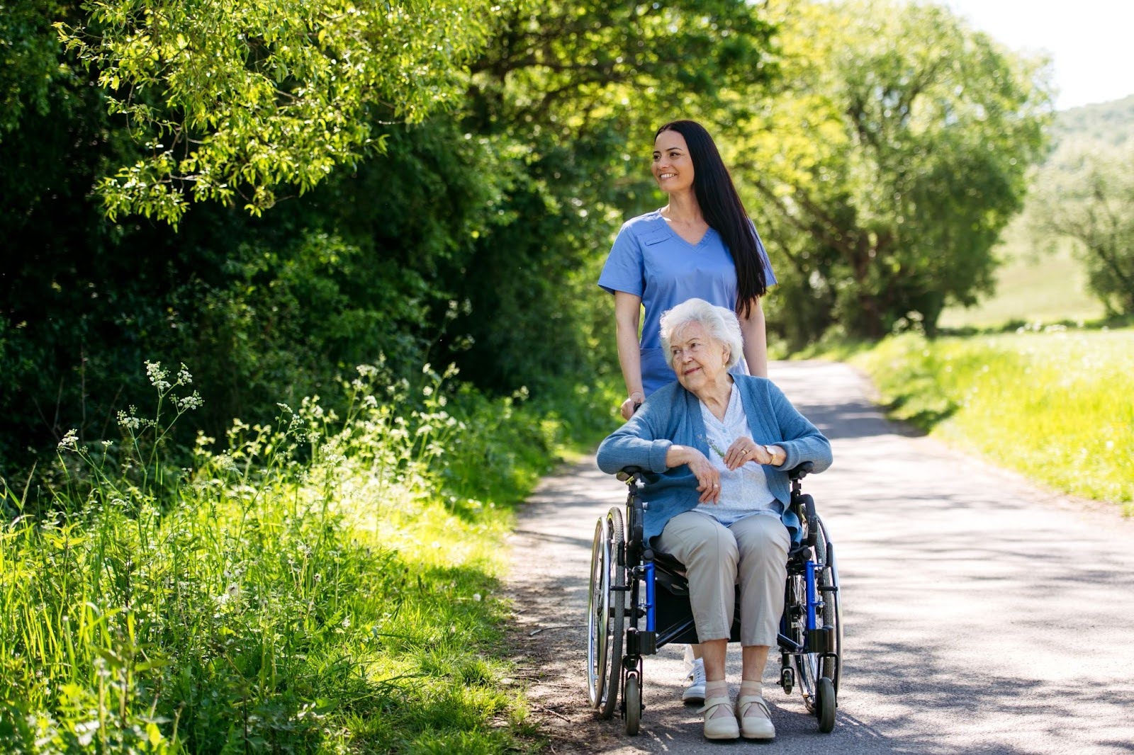 A caregiver pushing an older woman in a wheelchair along a tree-lined walking path.